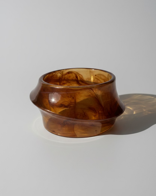 A translucent amber-brown glass bowl with a wide rim, resting on a beige striped textile with a reddish-brown border under direct sunlight.