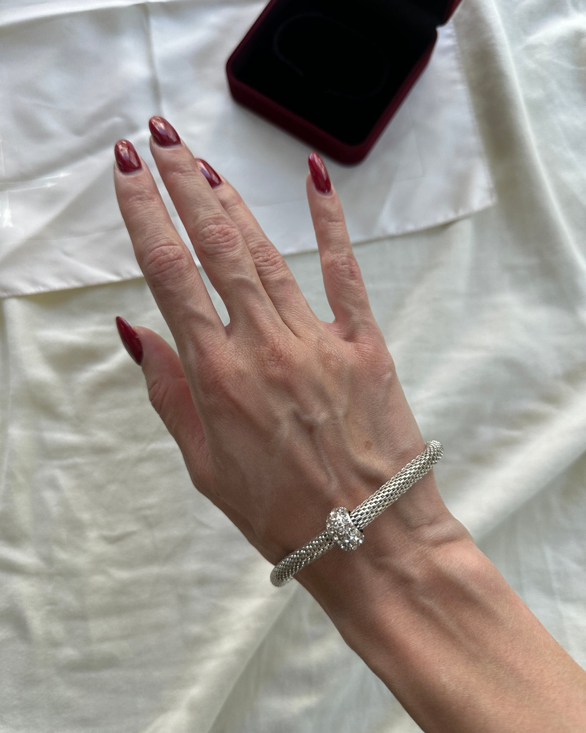 A close-up of a person's hand with dark red manicured nails wearing a silver braided bracelet with a central knot detail. A dark red jewelry box is partially visible on a white fabric background.