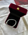 A silver-toned diamond-encrusted bracelet displayed inside an open dark red velvet jewelry box with a gold clasp, set on a white satin fabric.