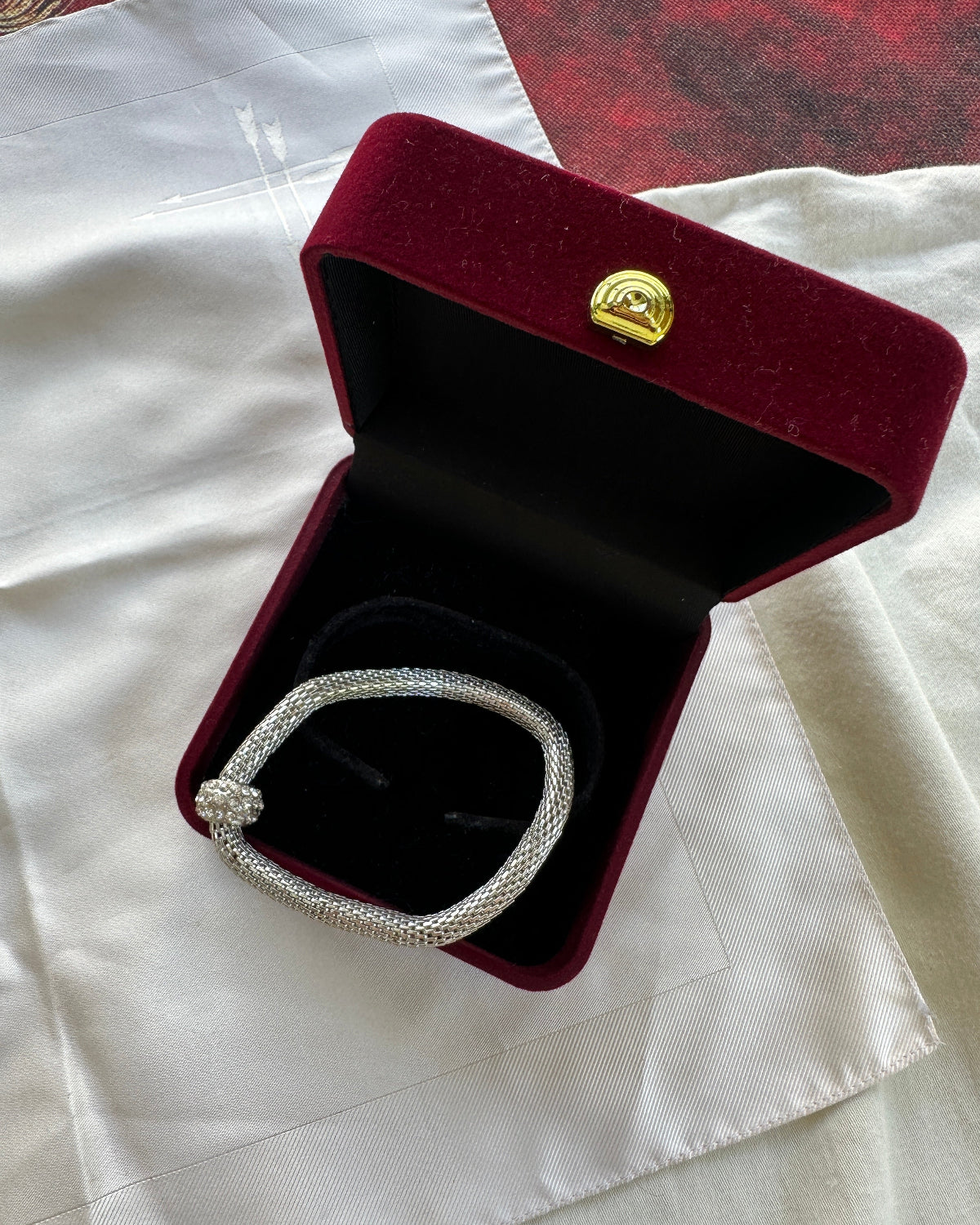 A silver-toned diamond-encrusted bracelet displayed inside an open dark red velvet jewelry box with a gold clasp, set on a white satin fabric.