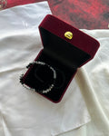 A beaded bracelet with black and clear beads displayed inside an open burgundy velvet jewelry box with a gold clasp, set on a white fabric surface.
