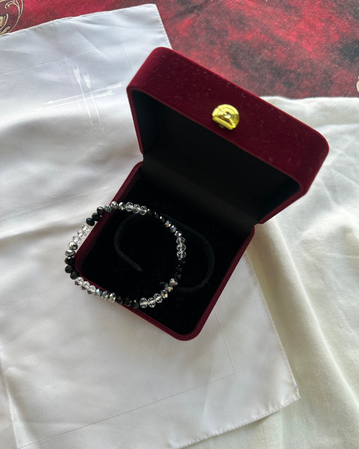 A beaded bracelet with black and clear beads displayed inside an open burgundy velvet jewelry box with a gold clasp, set on a white fabric surface.