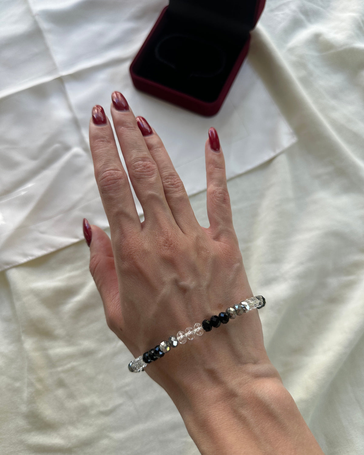 A close-up of a person's hand with dark red manicured nails wearing a beaded stretch bracelet. The bracelet features clear faceted beads, black beads, and silver-toned metal spacers.