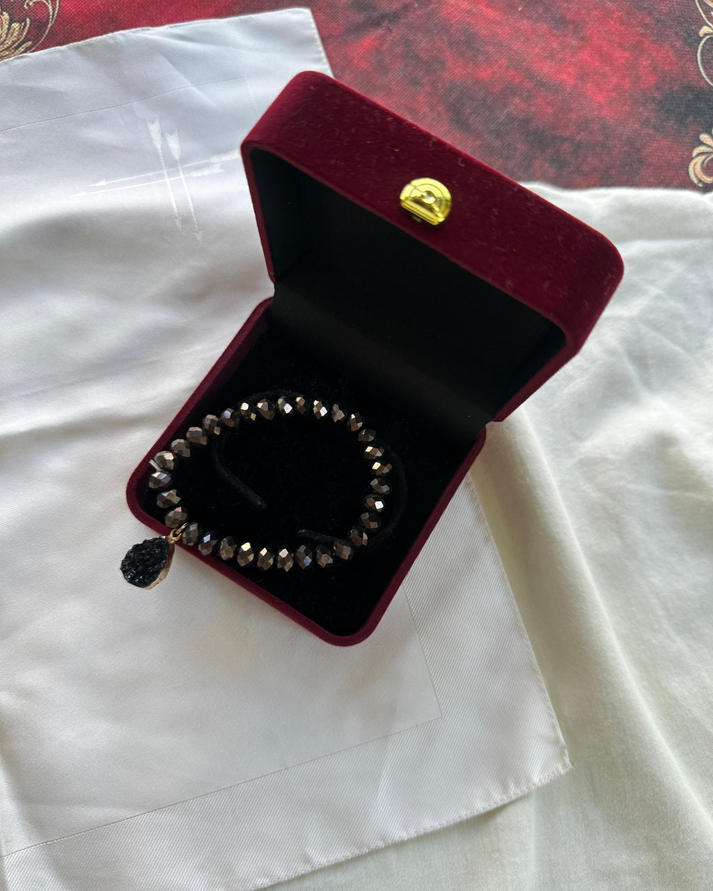A beaded bracelet with dark brown and black beads, featuring a small black tassel charm, presented inside an open dark red velvet jewelry box on a white fabric background.