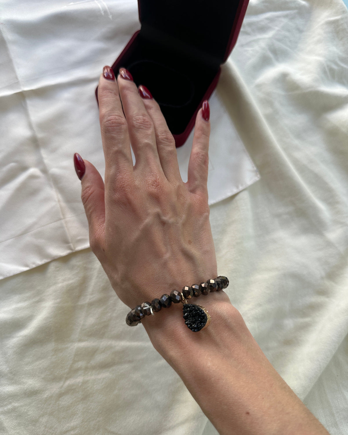 A close-up of a person's hand wearing a beaded bracelet with a black teardrop crystal pendant, resting near an open black jewelry box on a white fabric background.
