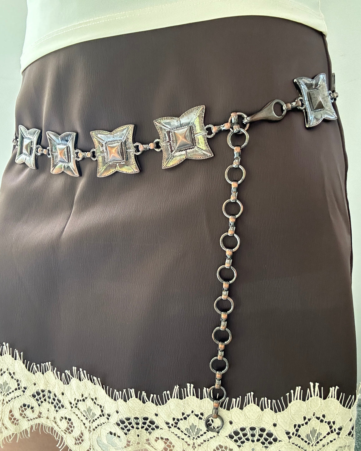 A brown satin mini skirt featuring a decorative silver chain belt with star-shaped crystal embellishments and a dangling chain, finished with a cream lace hem.