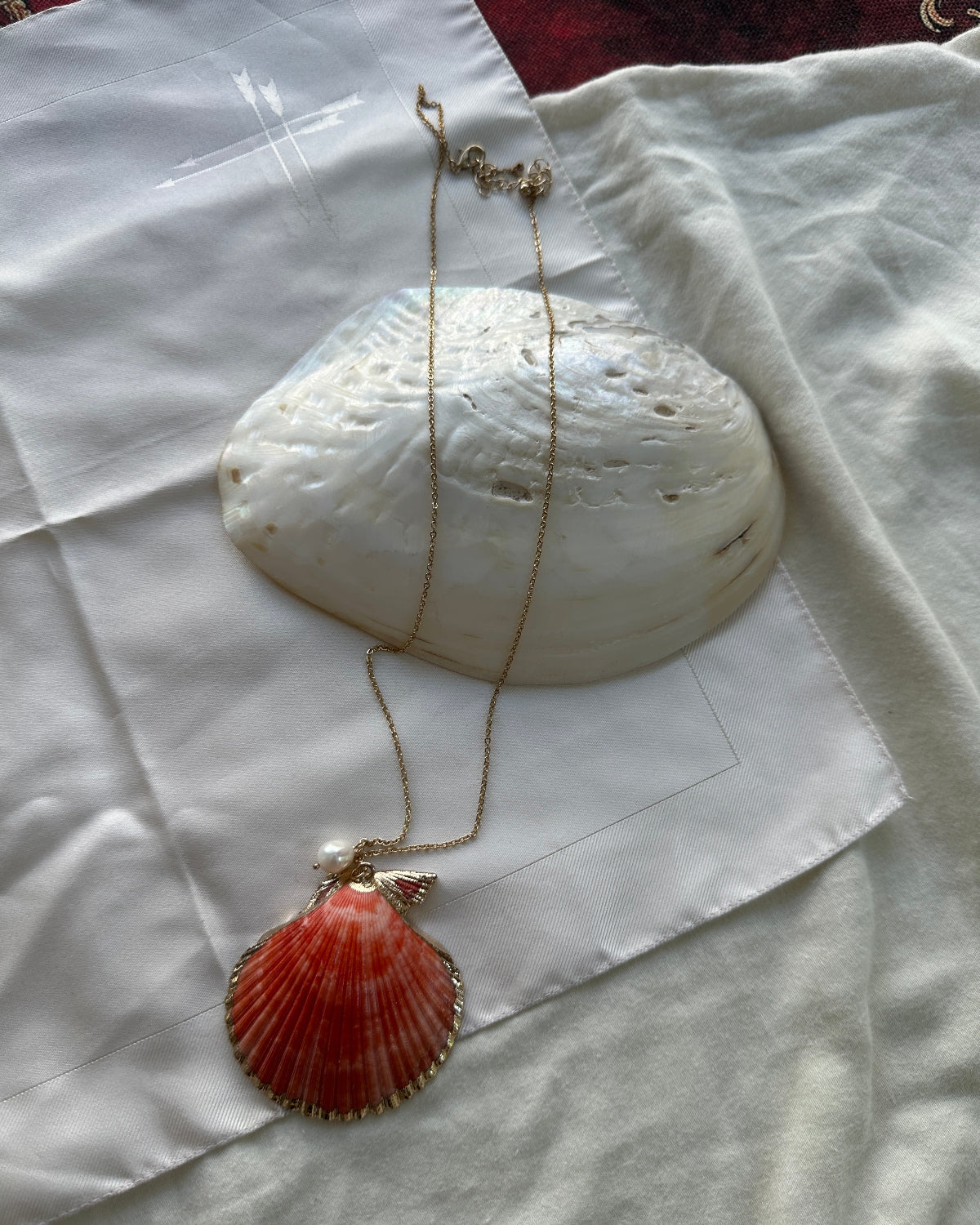 A gold chain necklace with a red-orange scallop shell pendant, displayed on a white fabric background with a larger white shell prop.