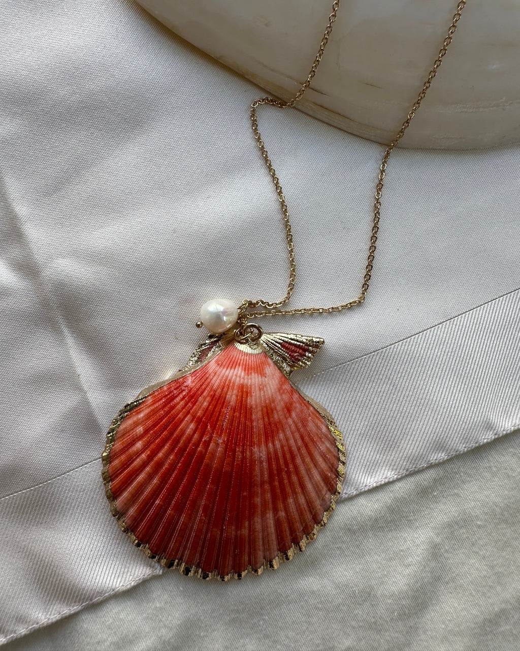 A gold-tone scallop shell pendant necklace with a small white pearl accent, displayed on a white satin ribbon next to a white stone.