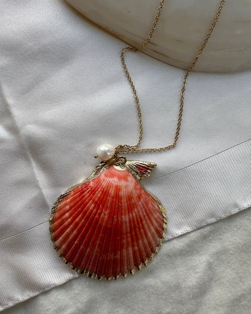 A gold-tone scallop shell pendant necklace with a small white pearl accent, displayed on a white satin ribbon next to a white stone.
