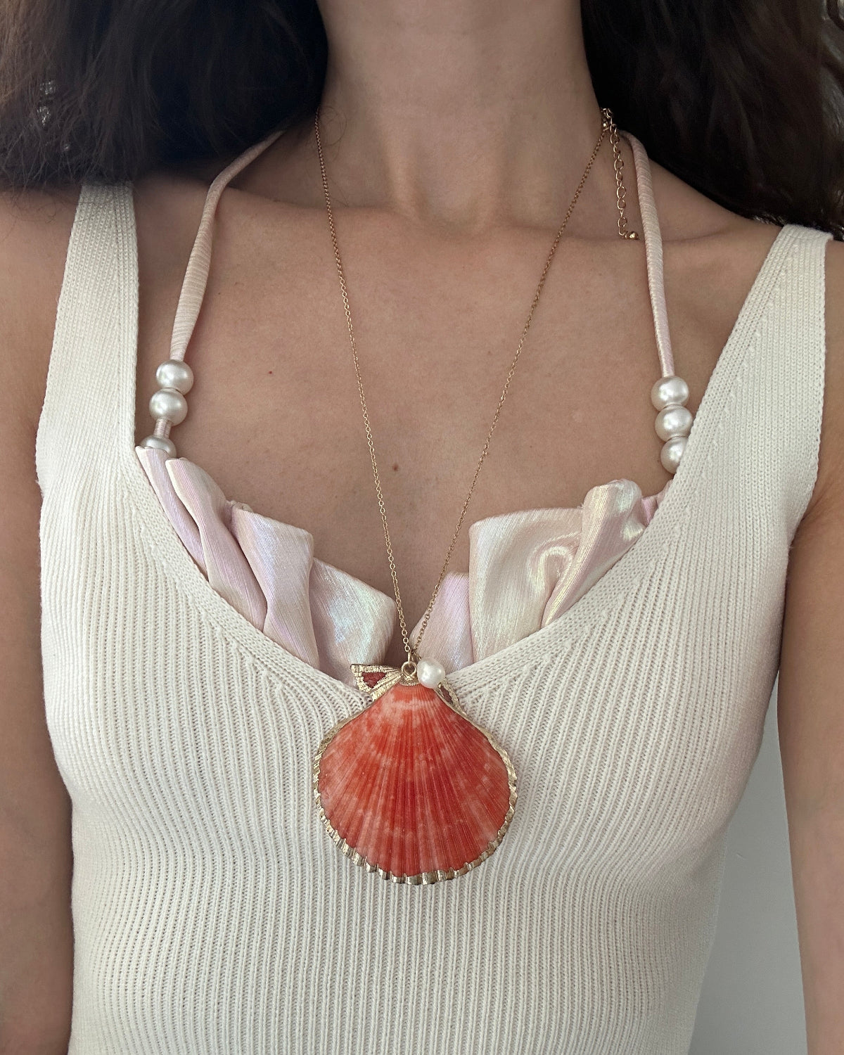 A close-up of a woman wearing a gold necklace with a large scallop shell pendant and pearl-beaded shoulder straps over a cream ribbed top.