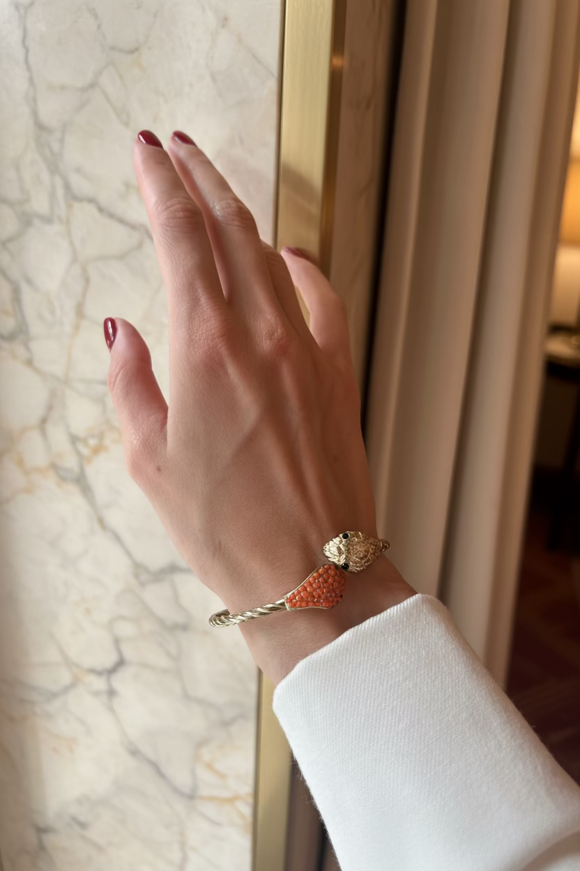 A close-up of a person's hand with red nail polish resting against a yellow curtain, wearing a silver-toned snake bracelet embellished with orange-red crystals.