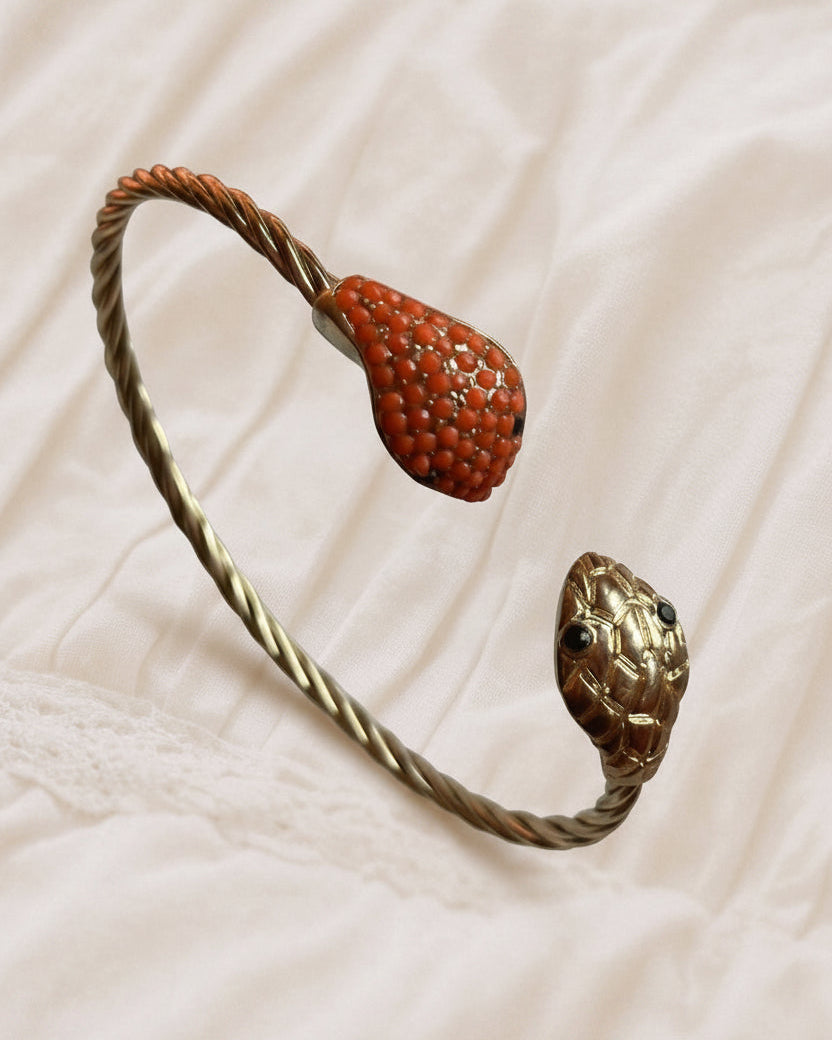 A twisted metal cuff bracelet featuring a textured red-orange teardrop pendant and a sculpted gold snake head, displayed on a soft beige fabric background.