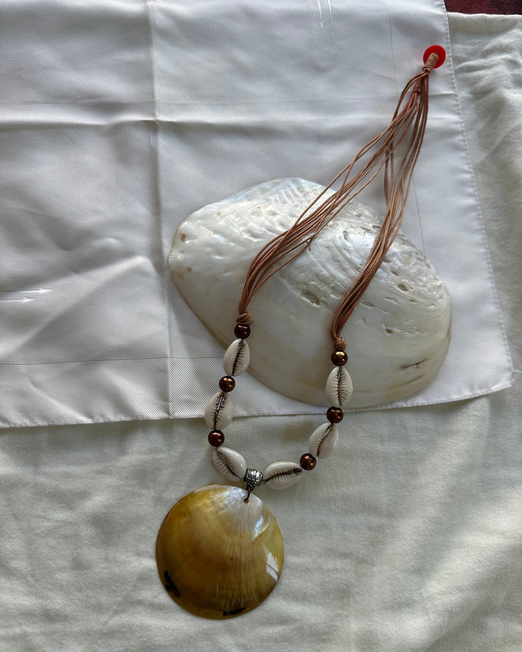 A handmade shell and bead necklace with a large oval yellow-brown shell pendant, displayed on a white fabric background with a larger white shell prop.