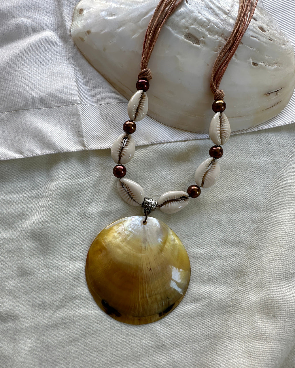 A bohemian-style shell necklace featuring a large iridescent yellow-brown clam shell pendant, white cowrie shells, dark brown beads, and a silver spacer, displayed on a white fabric background with a seashell prop.