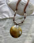 A bohemian-style shell necklace featuring a large iridescent yellow-brown clam shell pendant, white cowrie shells, dark brown beads, and a silver spacer, displayed on a white fabric background with a seashell prop.