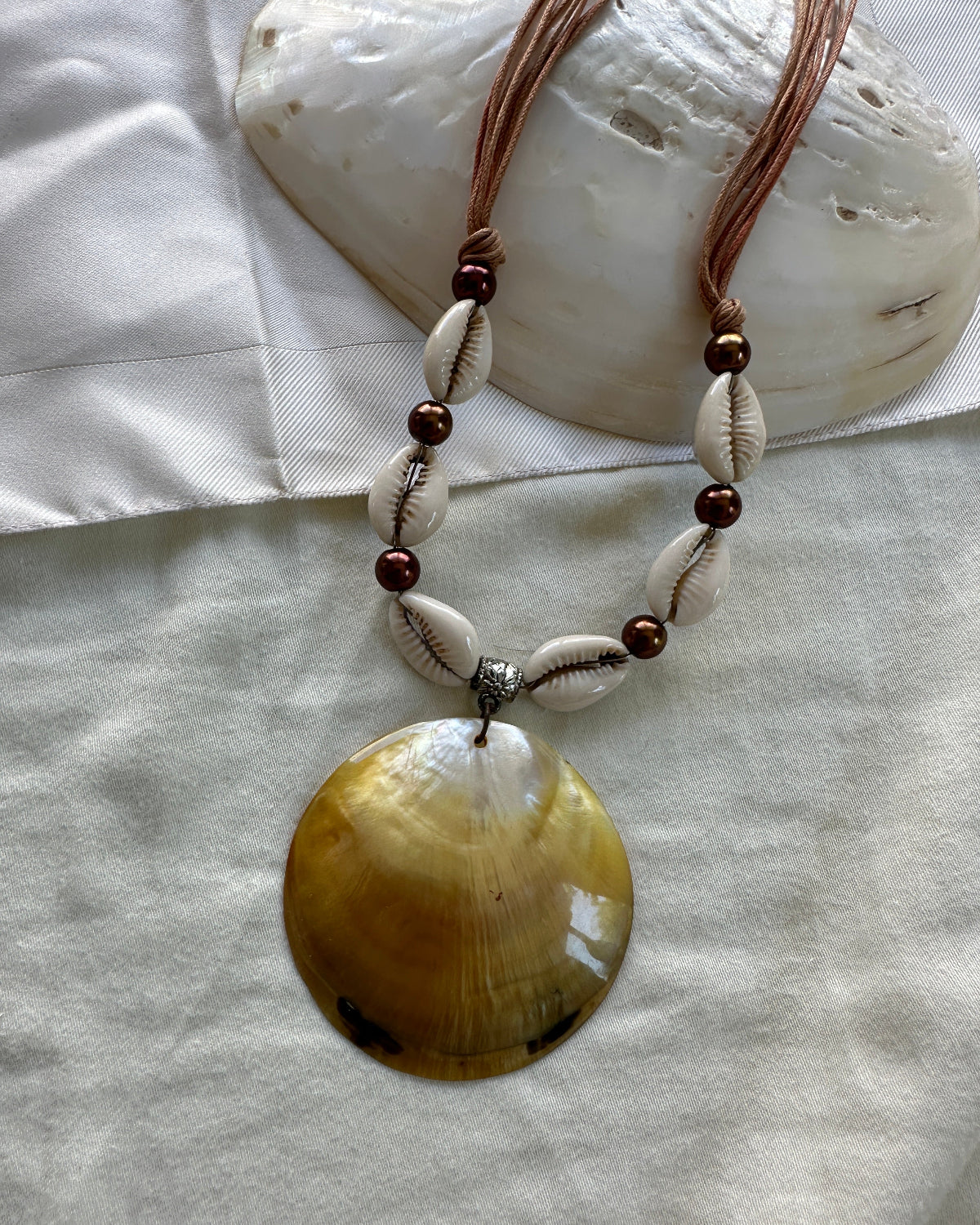 A bohemian-style shell necklace featuring a large iridescent yellow-brown clam shell pendant, white cowrie shells, dark brown beads, and a silver spacer, displayed on a white fabric background with a seashell prop.