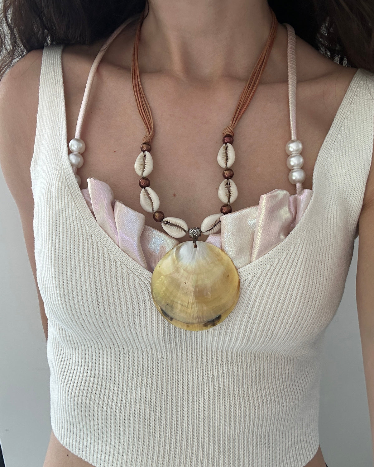 A close-up of a person wearing a cream ribbed tank top and a bohemian-style necklace with cowrie shells, brown beads, and a large yellow shell pendant.