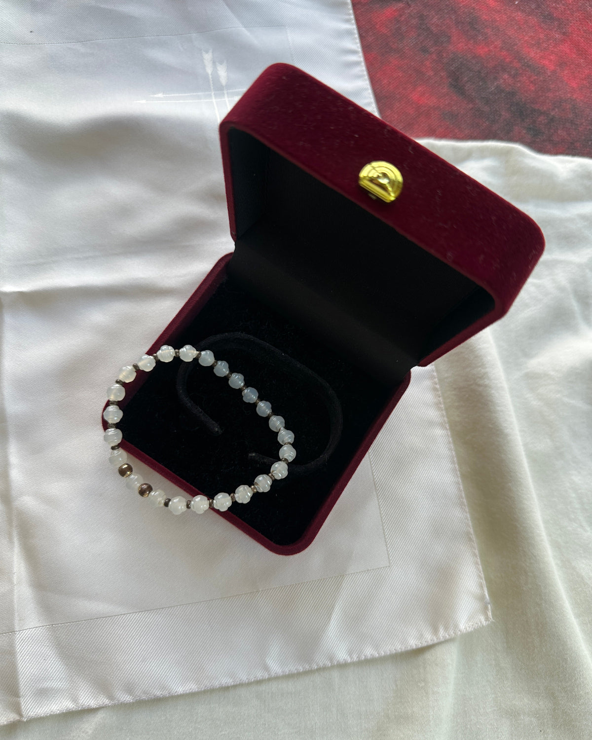 A beaded bracelet with white and silver beads displayed inside an open dark red velvet jewelry box with a gold clasp, set on a white satin fabric.