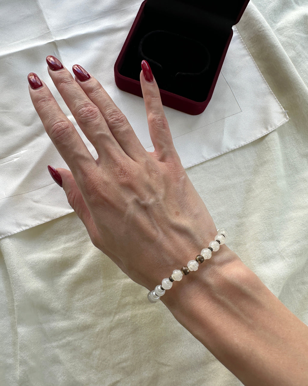 A close-up of a person's hand wearing a delicate beaded bracelet with white and clear faceted beads, resting on a cream-colored fabric surface next to an open dark red jewelry box.