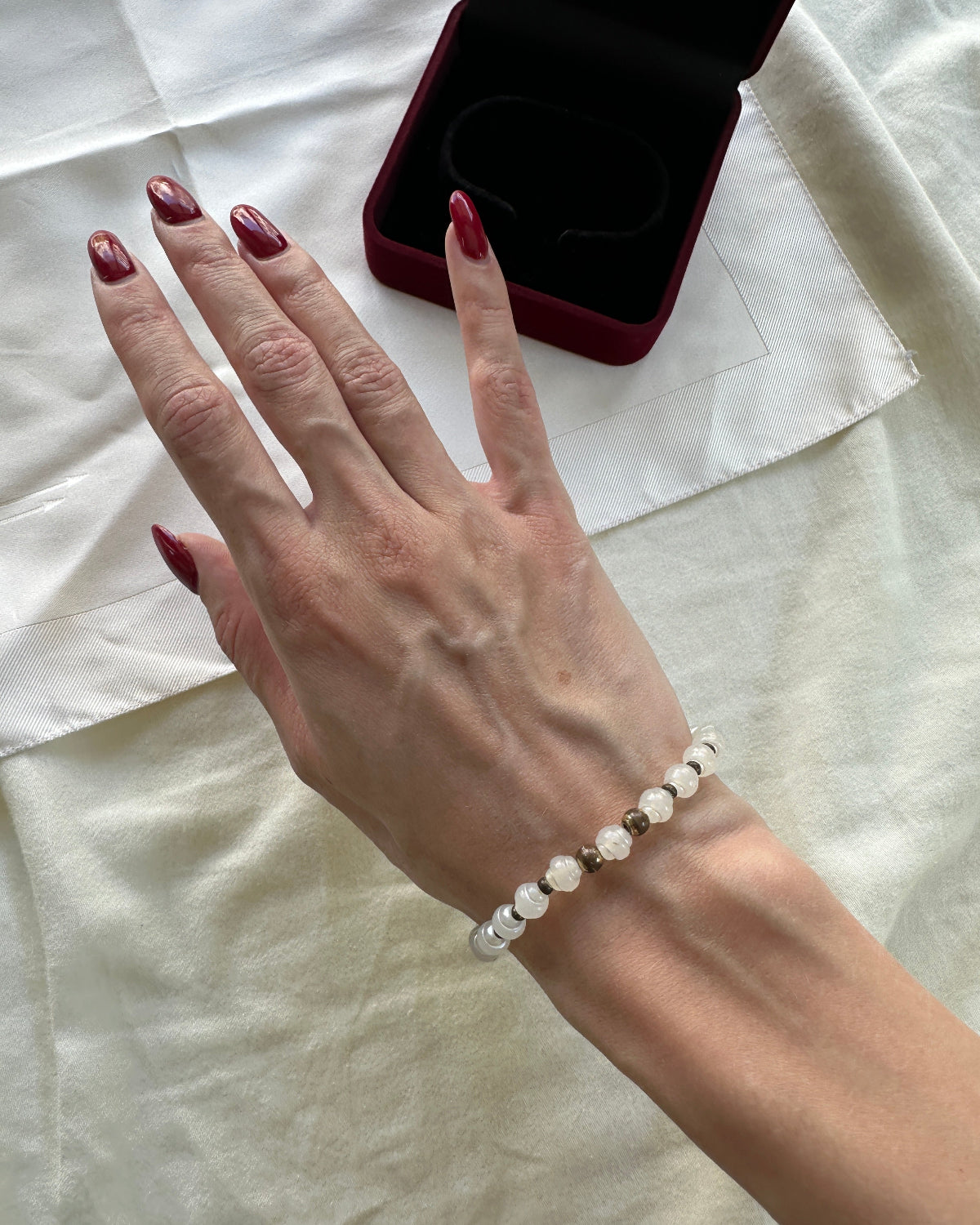 A close-up of a person's hand wearing a delicate beaded bracelet with white and clear faceted beads, resting on a cream-colored fabric surface next to an open dark red jewelry box.
