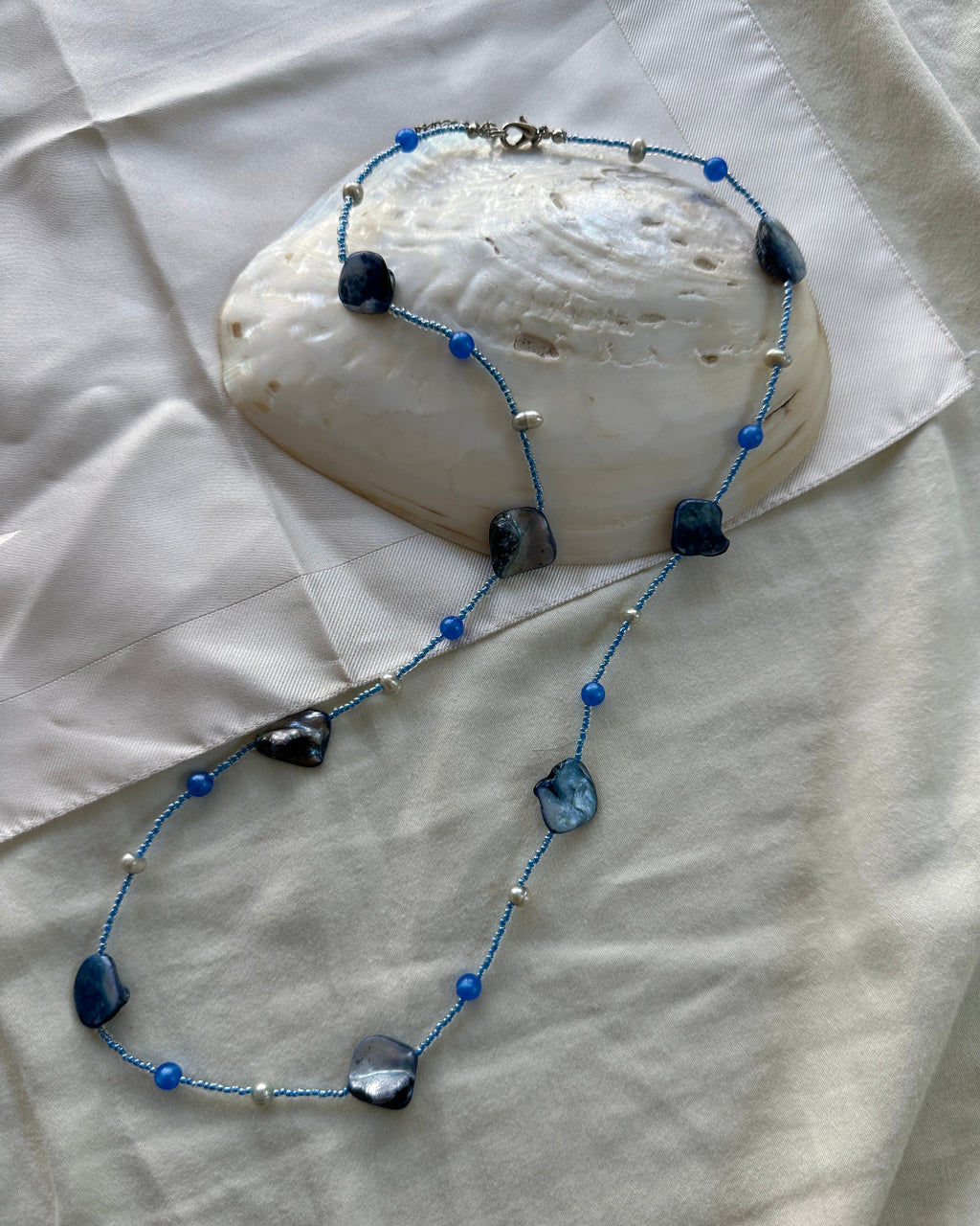 A beaded necklace with blue beads, irregular dark blue mother-of-pearl shell beads, and small silver spacer beads, displayed on a white fabric surface with a large white clam shell prop.