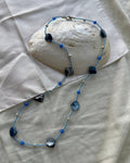 A beaded necklace with blue beads, irregular dark blue mother-of-pearl shell beads, and small silver spacer beads, displayed on a white fabric surface with a large white clam shell prop.