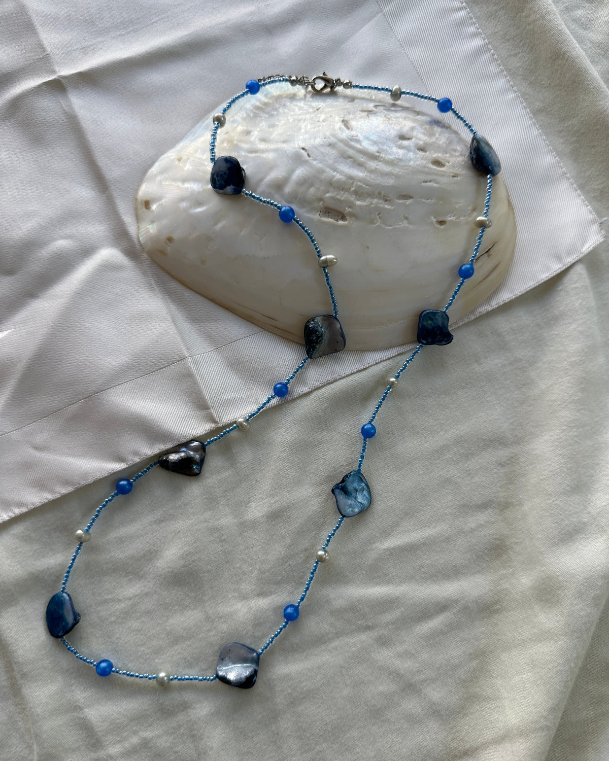 A beaded necklace with blue beads, irregular dark blue mother-of-pearl shell beads, and small silver spacer beads, displayed on a white fabric surface with a large white clam shell prop.