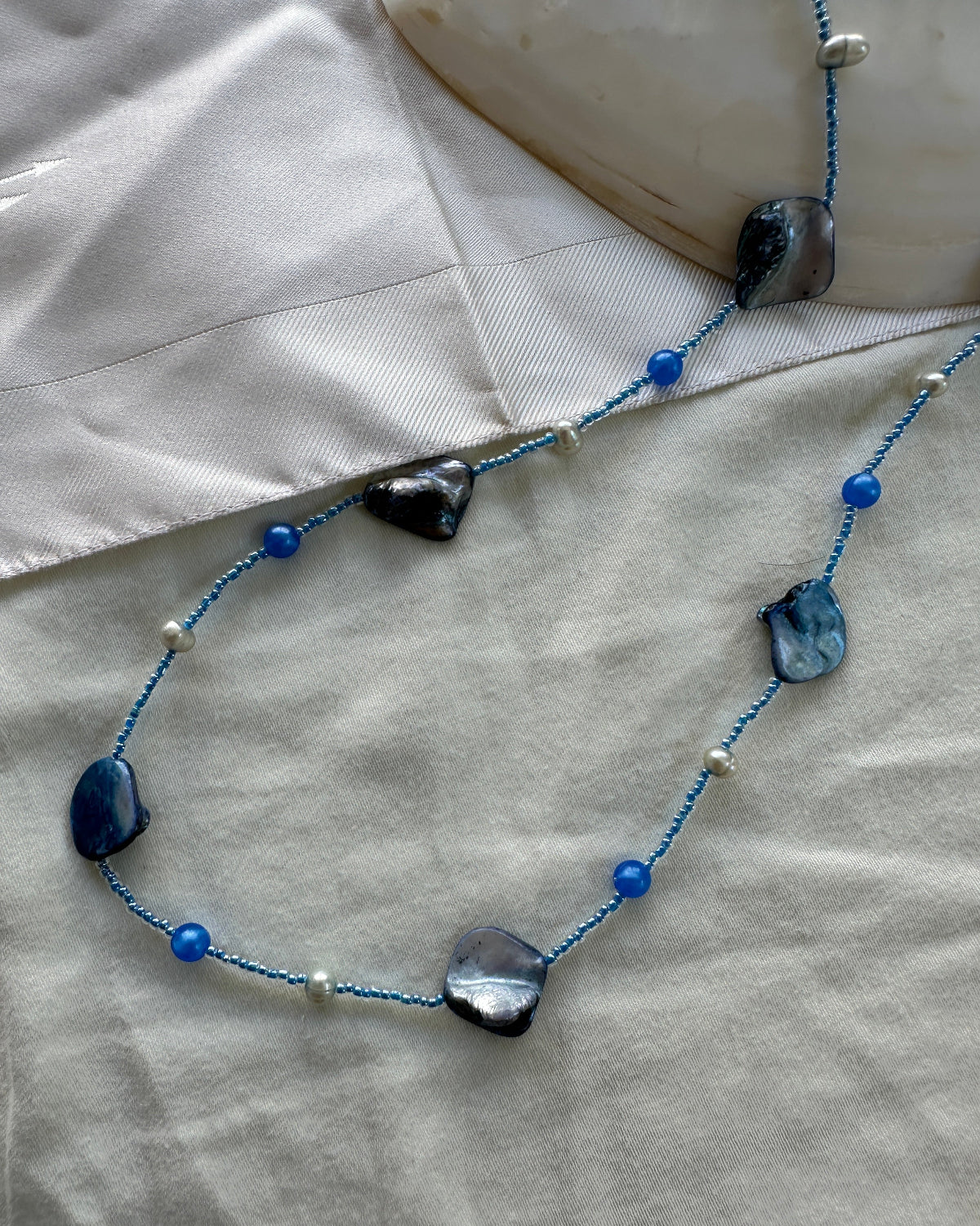 A beaded necklace with irregular dark blue and black stones, blue beads, and small white pearls on a blue cord, laid out on a light beige fabric surface.