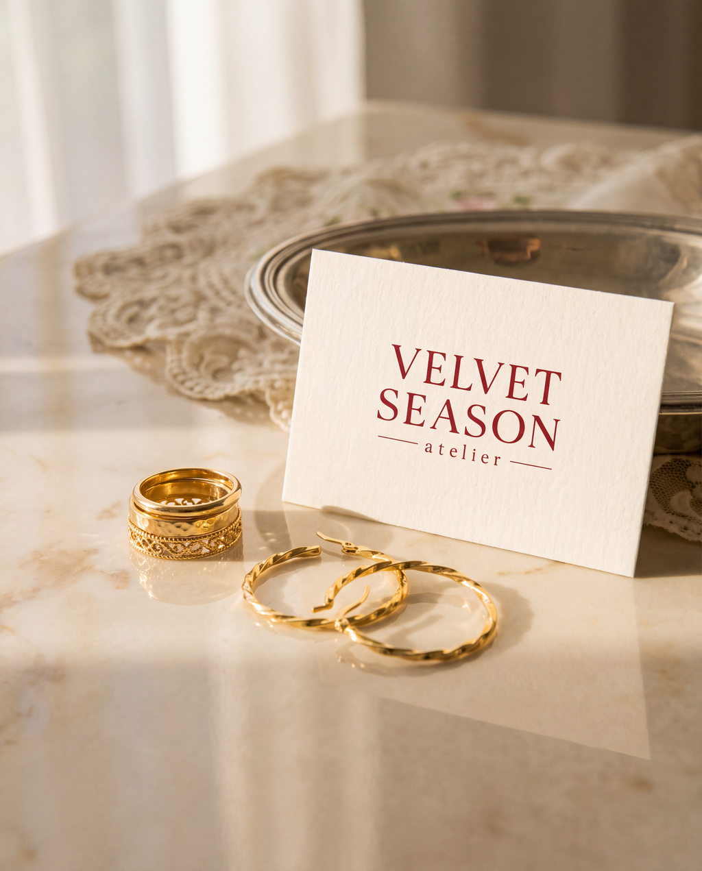 A styled flat-lay of gold jewelry on a marble surface, featuring a white card with the text 'VELVET SEASON atelier' in red. The jewelry includes textured gold hoop earrings and stacked gold rings, set against a background with a lace doily and a silver tray.
