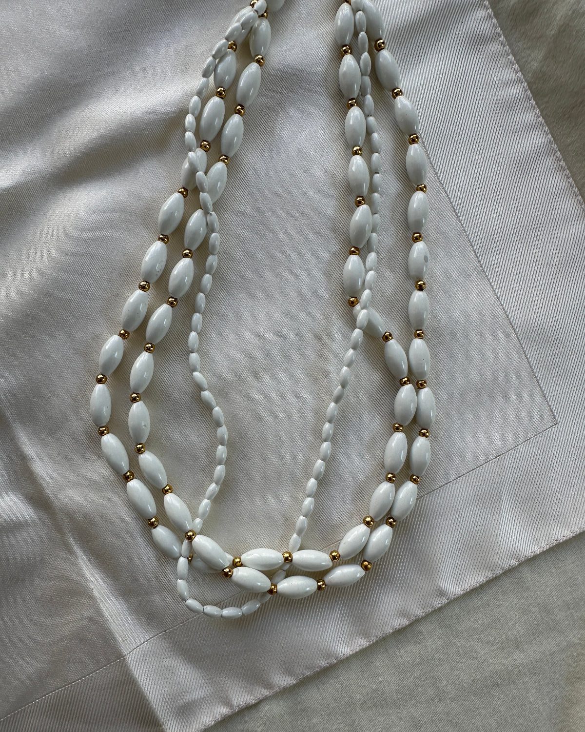 A multi-strand beaded necklace featuring white oval beads separated by small gold spacer beads, draped elegantly on a light beige textured fabric background.