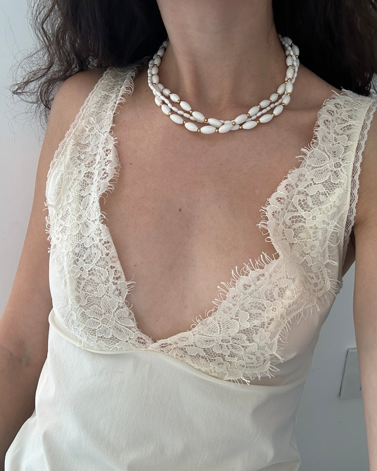 A close-up of a model wearing a cream-colored lace-trimmed camisole top and a multi-strand white shell necklace.