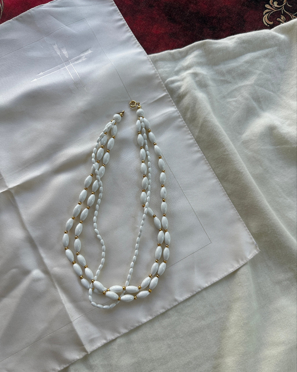 A white beaded necklace with gold-toned spacer details and a gold clasp, laid flat on a white satin fabric.