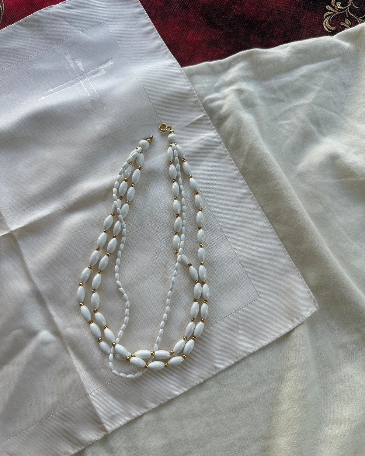 A white beaded necklace with gold-toned spacer details and a gold clasp, laid flat on a white satin fabric.