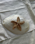 A textured gold starfish ornament resting on a large white seashell, placed on a white fabric background.