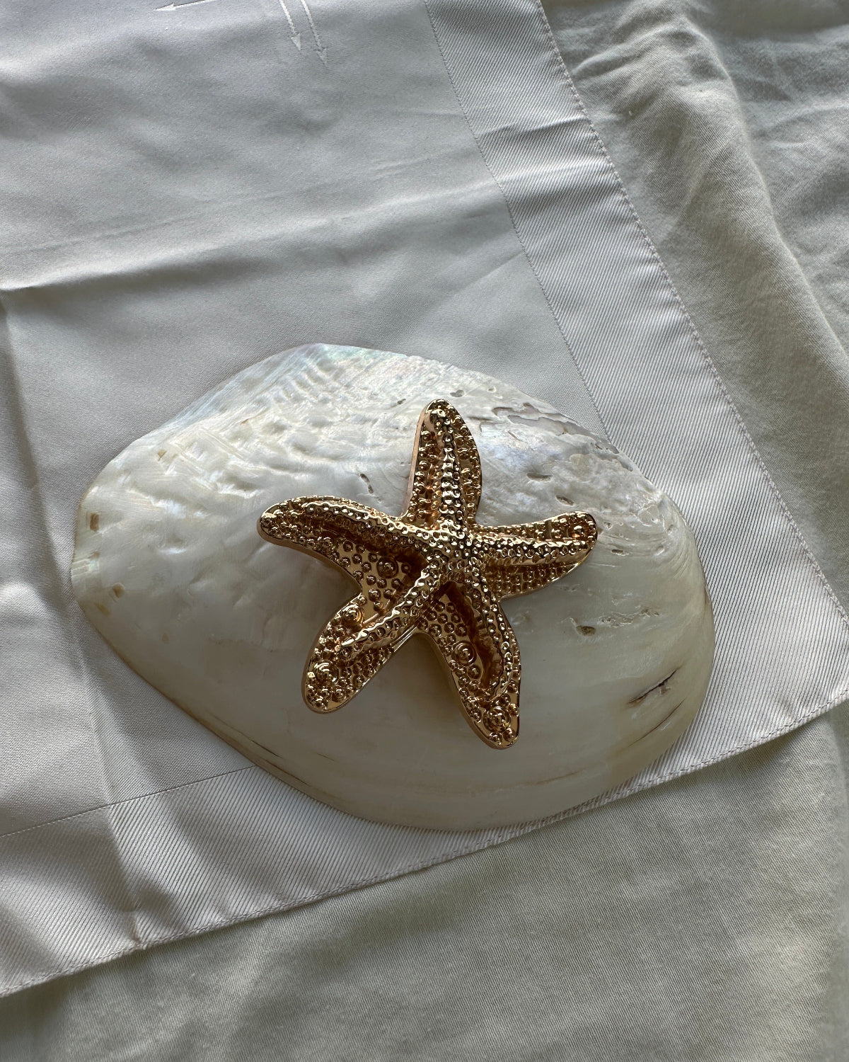 A textured gold starfish ornament resting on a large white seashell, placed on a white fabric background.