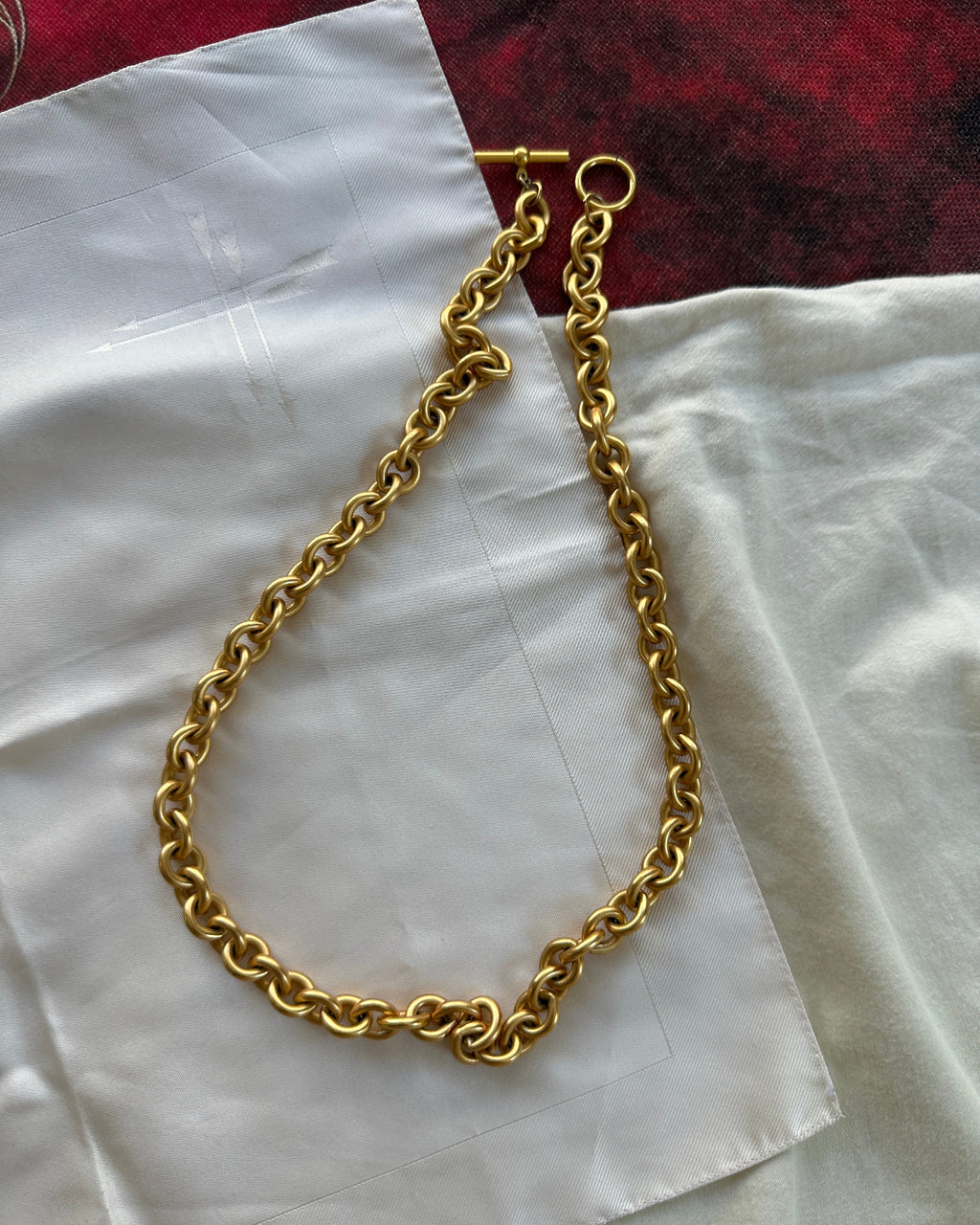 A chunky gold chain necklace with a toggle clasp, laid out on a white fabric surface with a red textured fabric in the background.