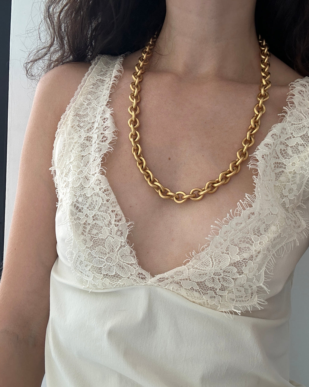 A close-up of a person wearing a chunky gold chain necklace over a cream lace-trim camisole top.