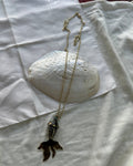 A gold chain necklace with a mermaid tail pendant, styled on a white fabric background with a large white clam shell prop.