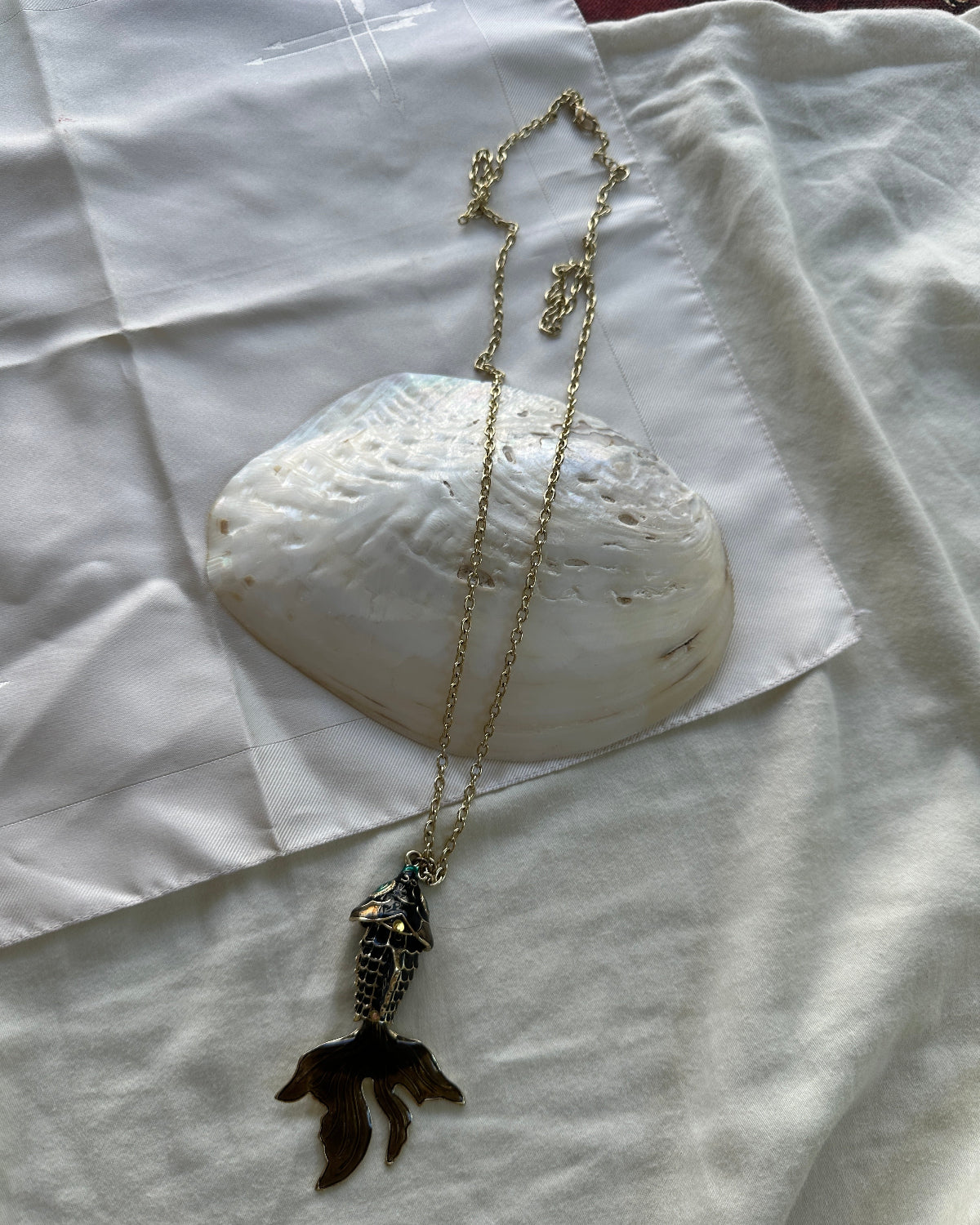 A gold chain necklace with a mermaid tail pendant, styled on a white fabric background with a large white clam shell prop.