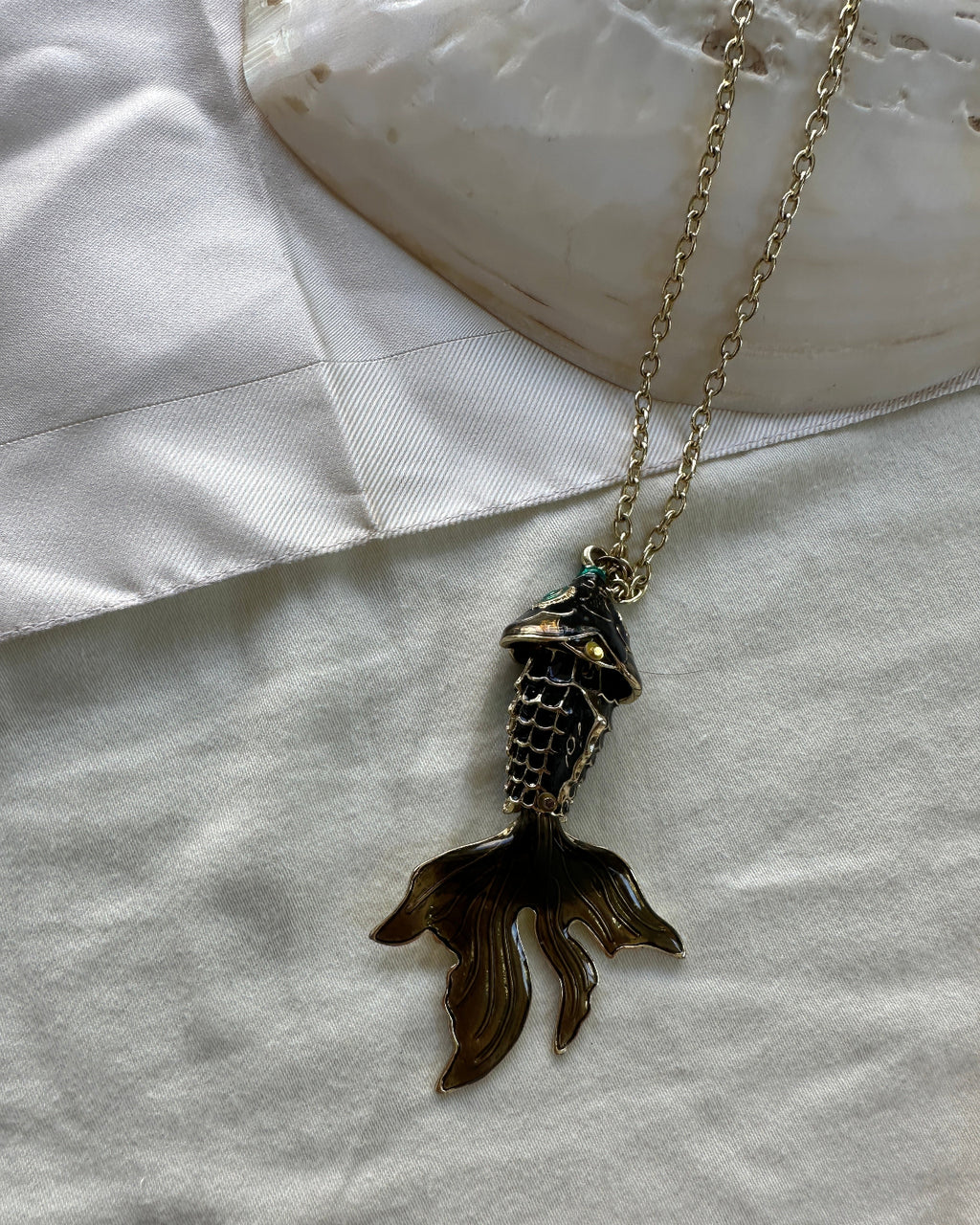 A gold-tone chain necklace with a mermaid tail pendant, styled on a white satin fabric next to a white shell.
