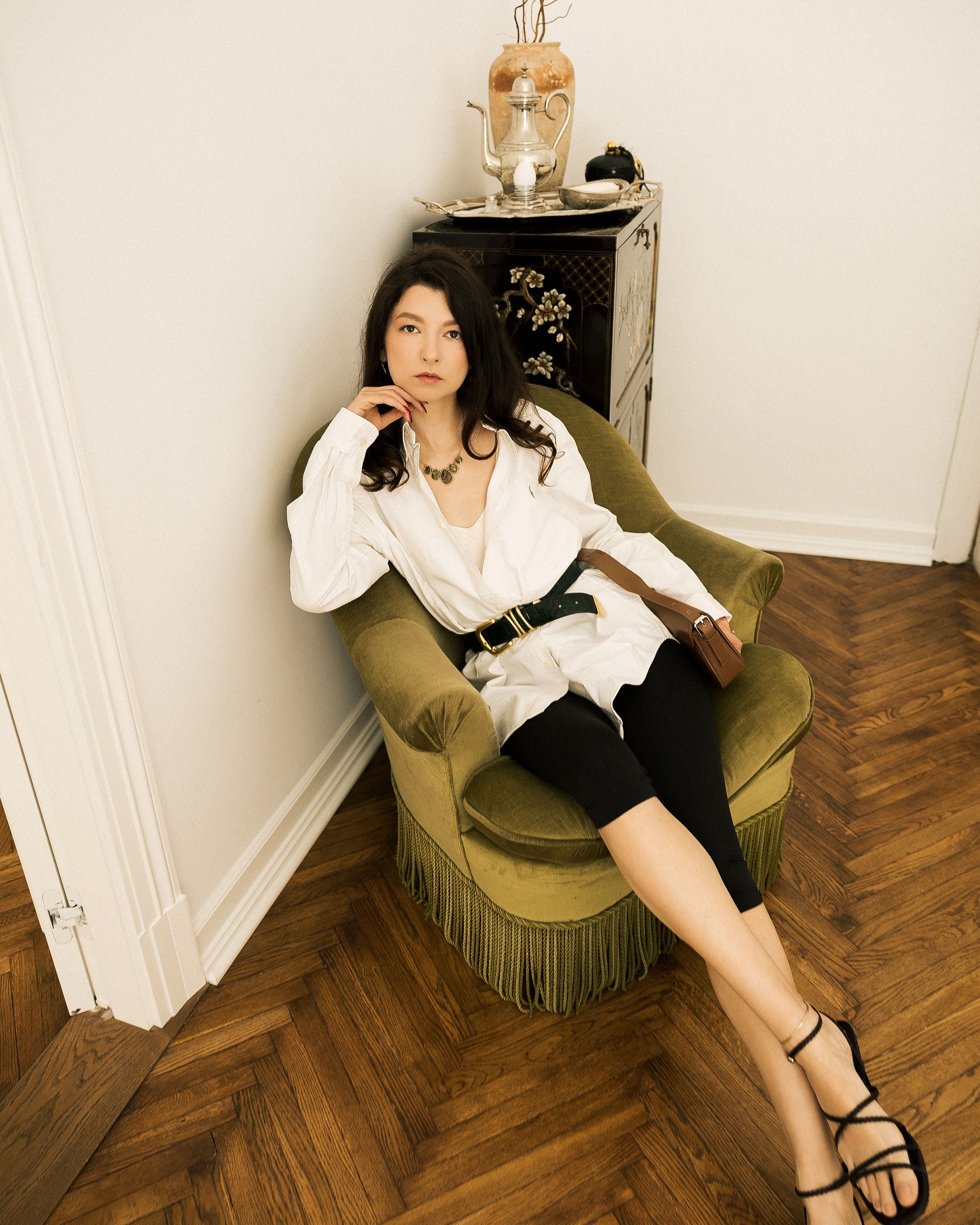 A woman wearing a white shirt, black skirt, and black strappy heels sits in a green fringed armchair in a room with a herringbone wood floor.