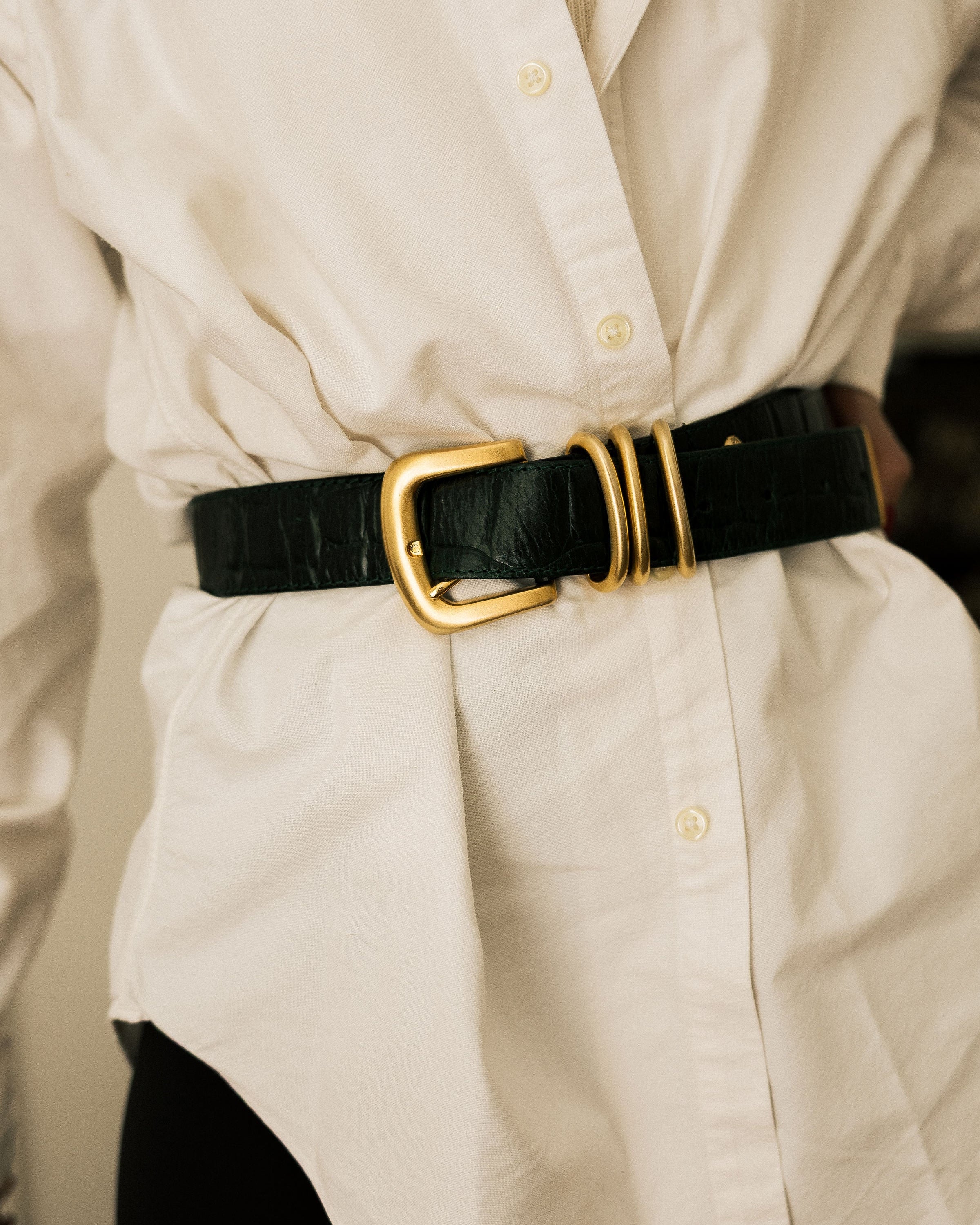 A close-up of a black crocodile-embossed belt worn at the waist over a cream-colored dress or shirt.