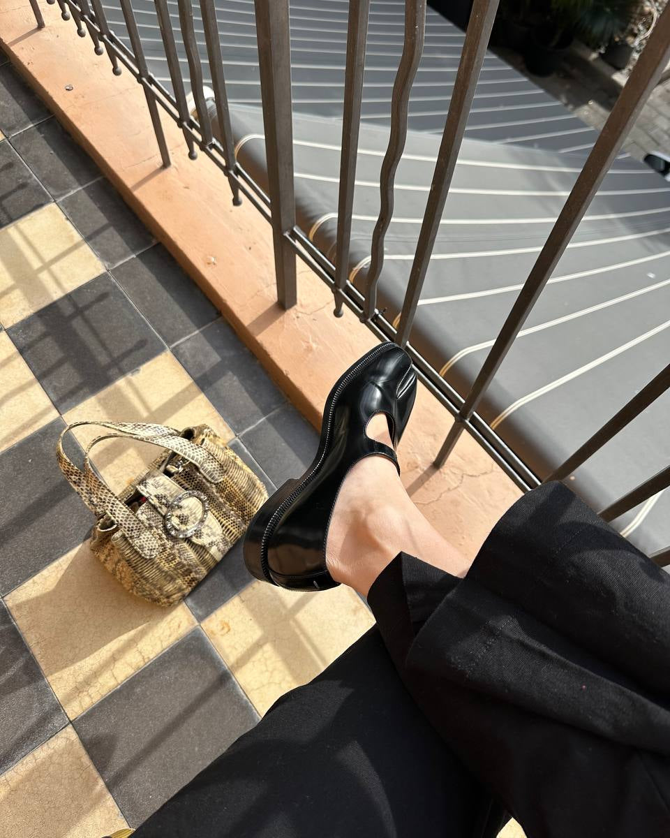A person wearing black leather Mary Jane flats with a chunky platform sole, photographed from a top-down perspective on a tiled balcony floor next to a snakeskin-patterned handbag.