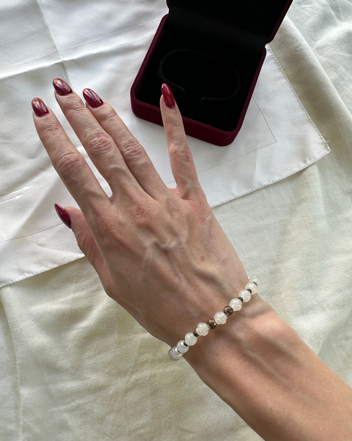 A close-up of a person's hand wearing a delicate beaded bracelet with white and clear faceted beads, resting on a cream-colored fabric surface next to an open dark red jewelry box.