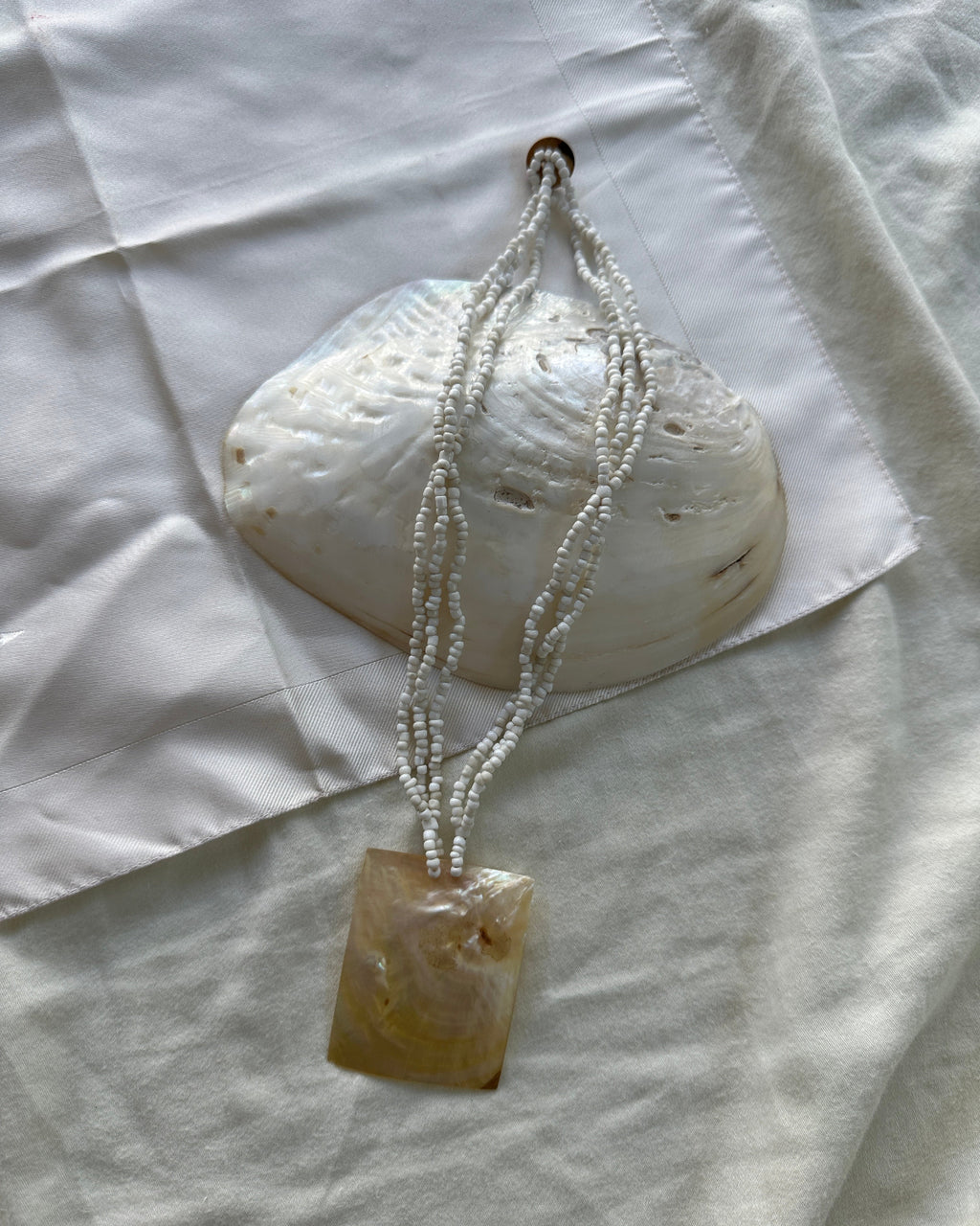 A handmade pendant necklace featuring a square, polished amber-brown stone pendant hanging from three strands of small white seed beads, displayed on a white fabric background with a white clam shell prop.