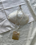 A handmade pendant necklace featuring a square, polished amber-brown stone pendant hanging from three strands of small white seed beads, displayed on a white fabric background with a white clam shell prop.