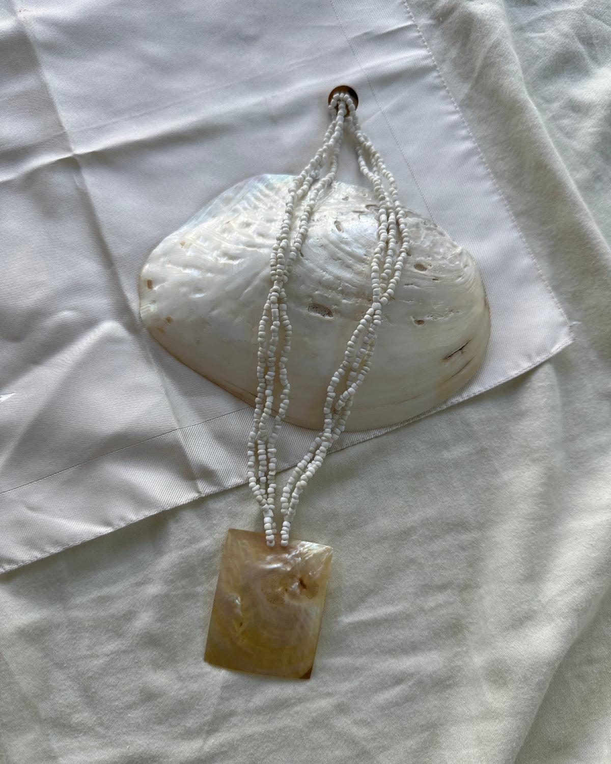 A handmade pendant necklace featuring a square, polished amber-brown stone pendant hanging from three strands of small white seed beads, displayed on a white fabric background with a white clam shell prop.
