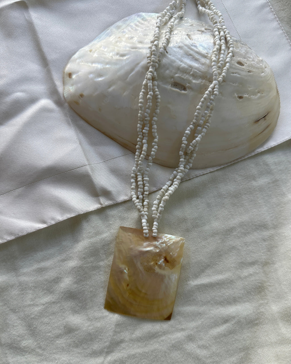 A beaded necklace with a rectangular mother-of-pearl pendant, displayed on a white fabric surface next to a larger shell.