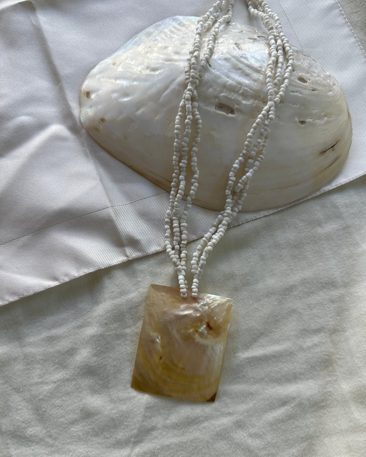 A beaded necklace with a rectangular mother-of-pearl pendant, displayed on a white fabric surface next to a larger shell.