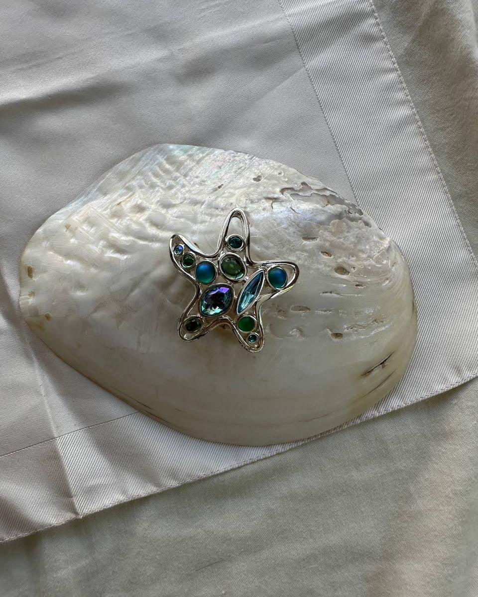 A silver starfish-shaped brooch with green and blue iridescent rhinestones, displayed on a large white clam shell over a cream-colored fabric background.